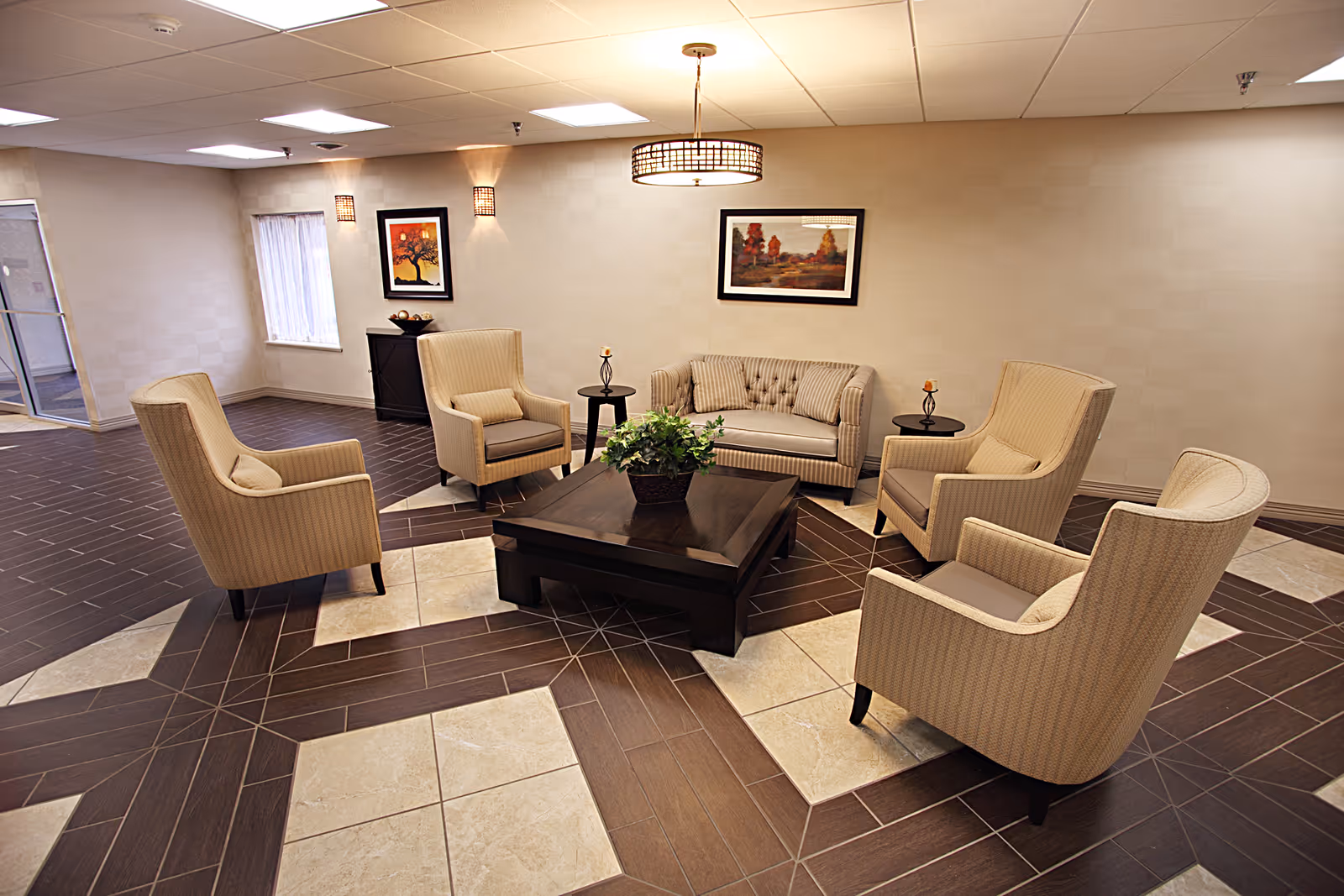 A cozy seating area in a senior living facility with a beige sofa and four matching armchairs arranged around a dark wooden coffee table with a plant centerpiece. The room has a patterned tile floor, two framed paintings on the wall, and soft lighting from ceiling fixtures and wall sconces.