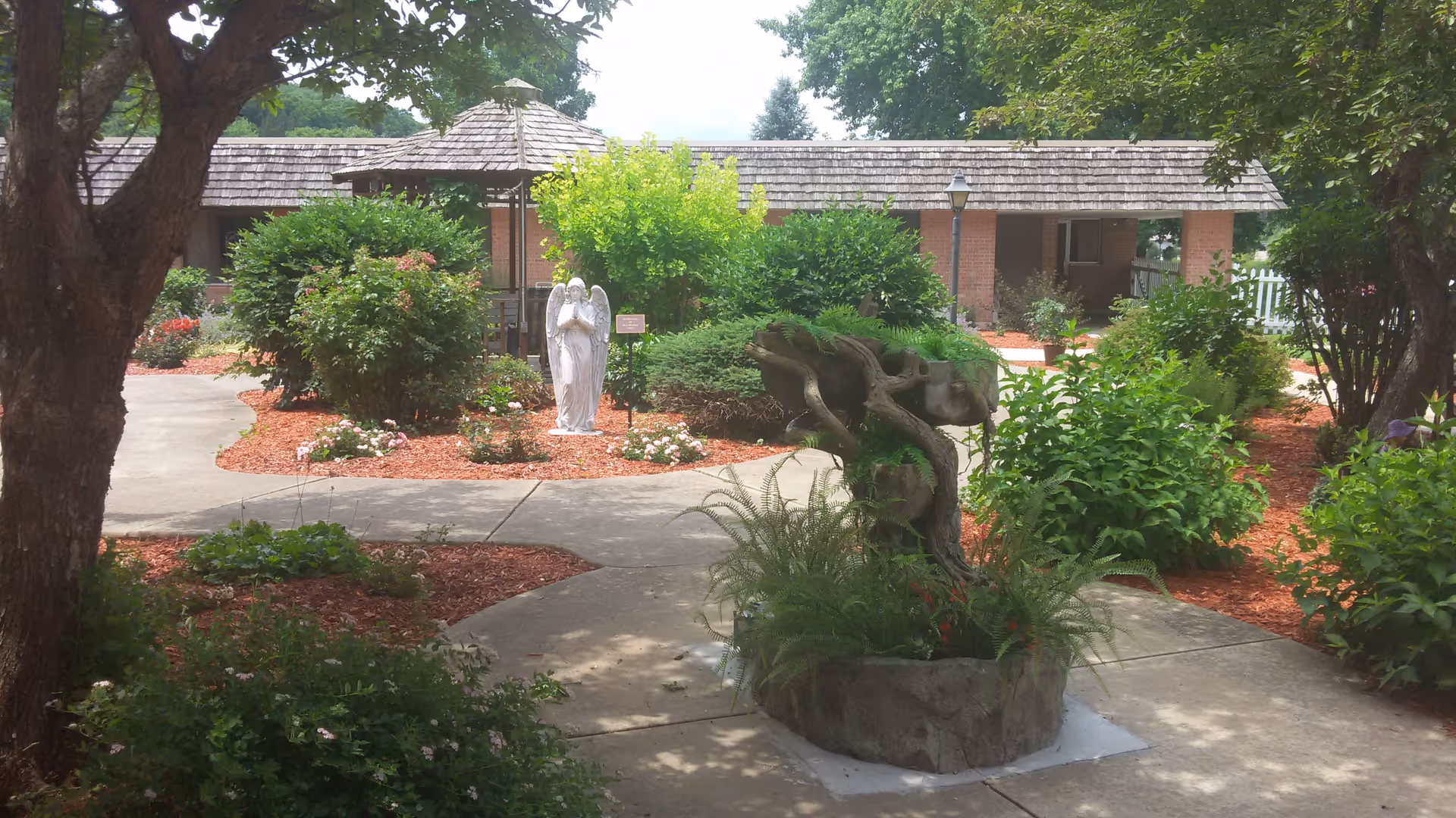 A peaceful outdoor garden area at Schmitt Woodland Hills Retirement Community featuring a concrete pathway, lush green bushes, trees, a white angel statue, a wooden gazebo with a shingled roof, and a decorative stone planter with ferns.