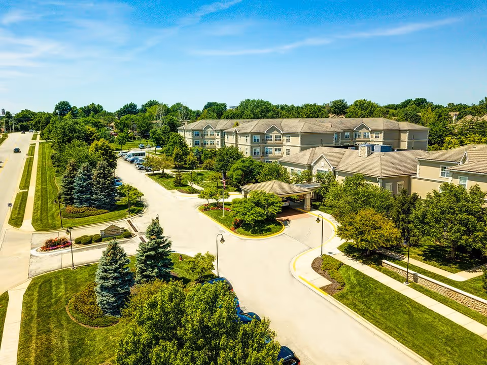 Aerial view of Town Village of Leawood senior living facility showing multiple beige buildings with gray roofs surrounded by green trees and landscaped lawns. There is a driveway with parked cars and a covered entrance area. The sky is clear and blue.