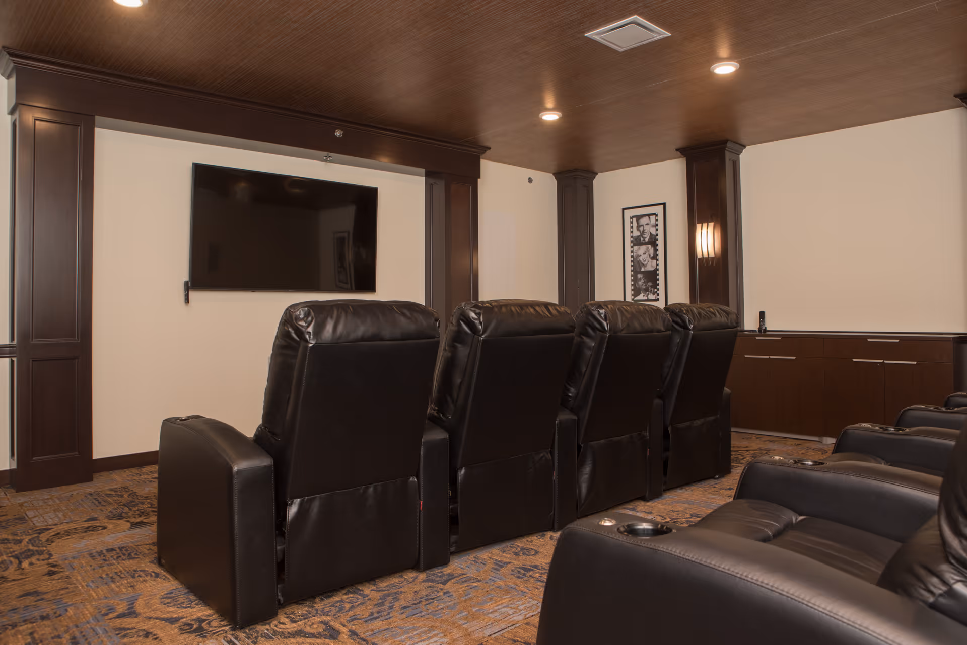 A cozy media room with two rows of black leather recliner chairs facing a large flat-screen TV mounted on a cream-colored wall. The room features dark wood paneling, a patterned carpet, recessed ceiling lights, and a framed black and white photo on the wall.