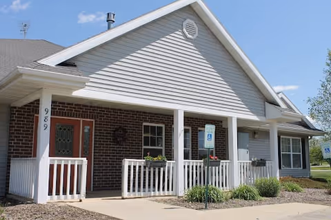 Exterior view of a single-story building with a covered porch, white railing, and brick and siding facade under a blue sky with some clouds. There are two handicap parking signs visible near the sidewalk leading to the entrance.