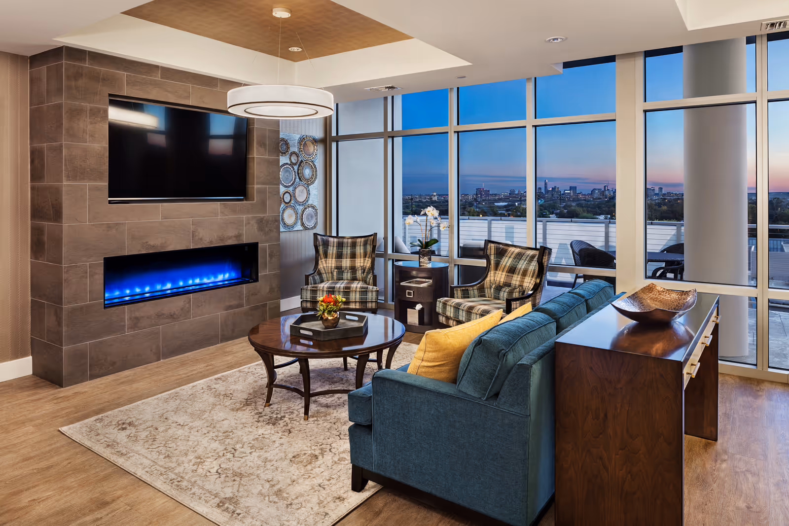 A modern living room with a blue sofa, two plaid armchairs, a round wooden coffee table with a flower arrangement, a wall-mounted flat-screen TV above a blue-lit electric fireplace, large floor-to-ceiling windows showing a cityscape at dusk, and a wooden console table with a decorative bowl.