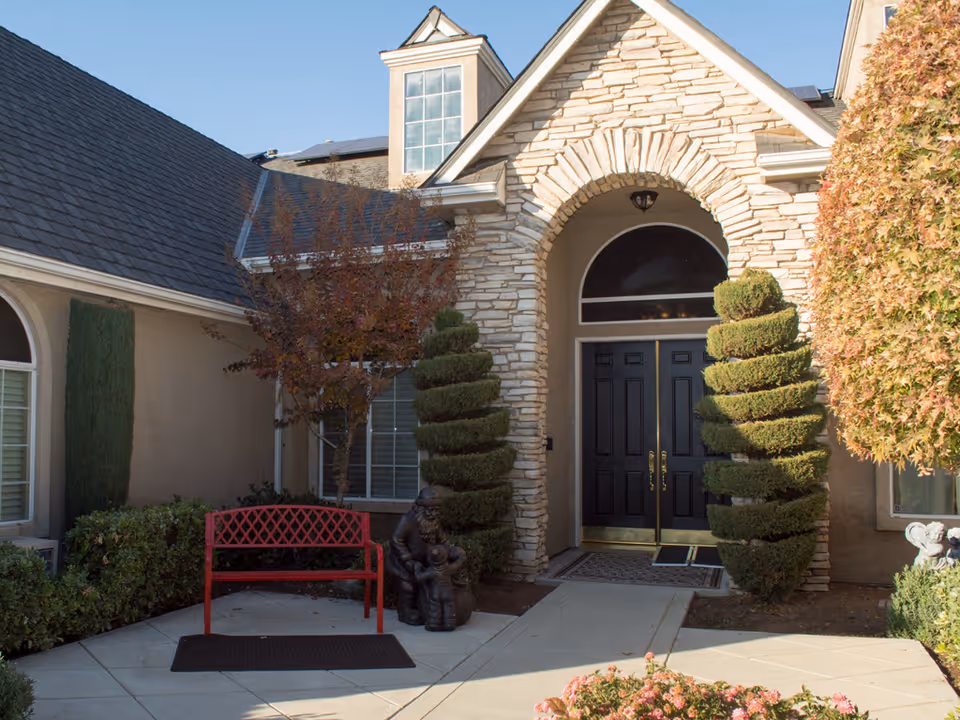 Entrance of a residential building with a stone archway and double black doors. There are two spiral-shaped topiary bushes on either side of the entrance, a red bench to the left, and a small statue of a person with children near the bench. The area is landscaped with bushes and trees, and the sky is clear and blue.