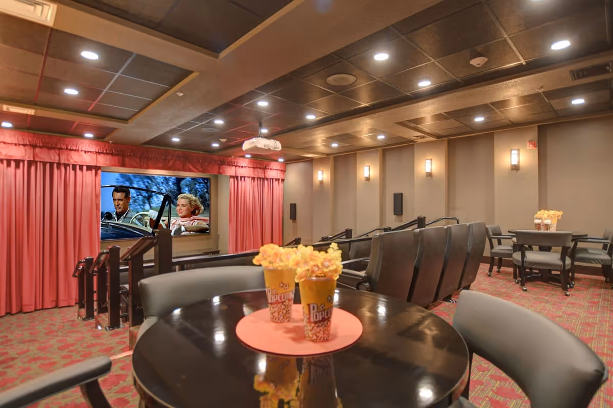 Interior view of a small movie theater room with a large screen showing a classic film. The room has red curtains around the screen, rows of black theater seats, and round tables with chairs. On the tables are containers of popcorn. The ceiling has recessed lighting and the walls are decorated with sconces.