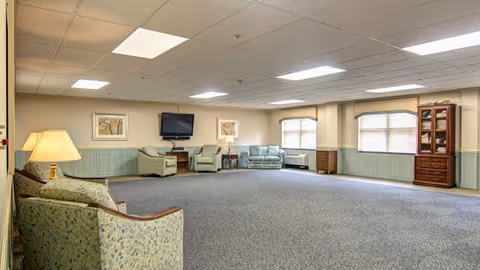 A spacious common area with carpeted floor, several upholstered armchairs and a sofa arranged along the walls, a wall-mounted flat screen TV, framed artwork, two windows with blinds, a wooden cabinet, and a table lamp on a side table.