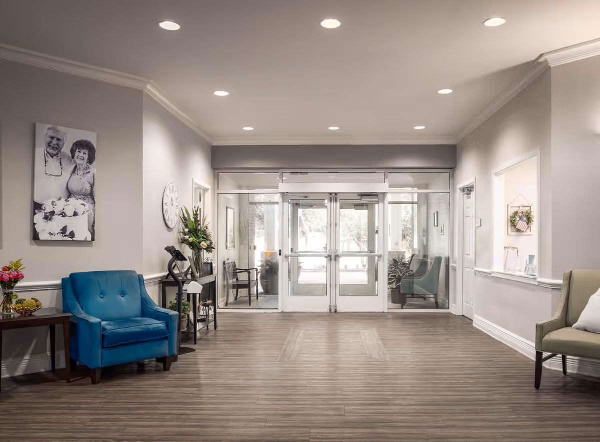 A spacious and well-lit senior living facility lobby with wood flooring, a blue armchair on the left, a green armchair on the right, a small table with flowers, a wall clock, and a black and white photo of an elderly couple on the left wall. The entrance features double glass doors with windows on either side.