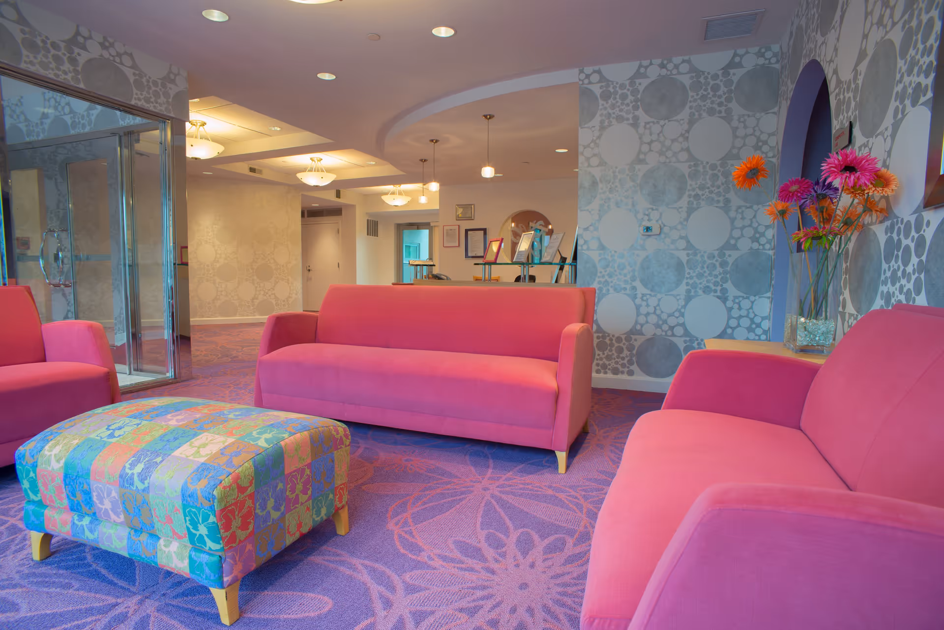 A brightly colored lounge area with three pink sofas arranged around a multicolored patterned ottoman on a purple carpet with floral designs. The walls have a circular pattern wallpaper, and there is a glass door on the left side. A vase with colorful flowers is placed on a side table to the right.