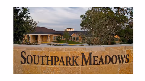 Stone sign with the text 'Southpark Meadows' in front of a single-story building surrounded by trees and greenery under a cloudy sky.