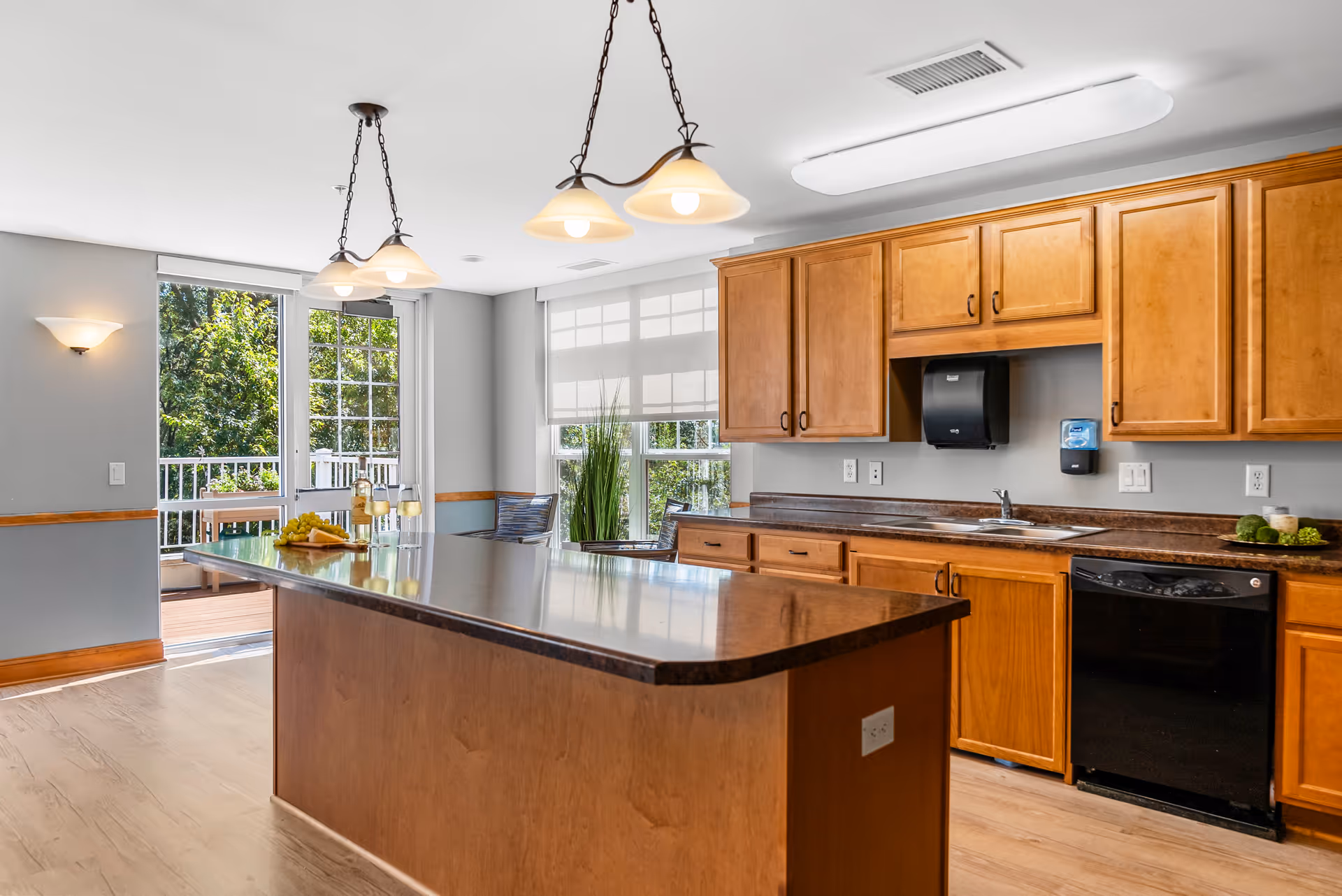 A bright kitchen area with wooden cabinets and a large island countertop in the center. The kitchen features a black dishwasher, a sink, and a paper towel dispenser mounted on the wall. Two pendant lights hang above the island. There is a window with a white blind and a door leading to an outdoor deck with greenery visible outside.