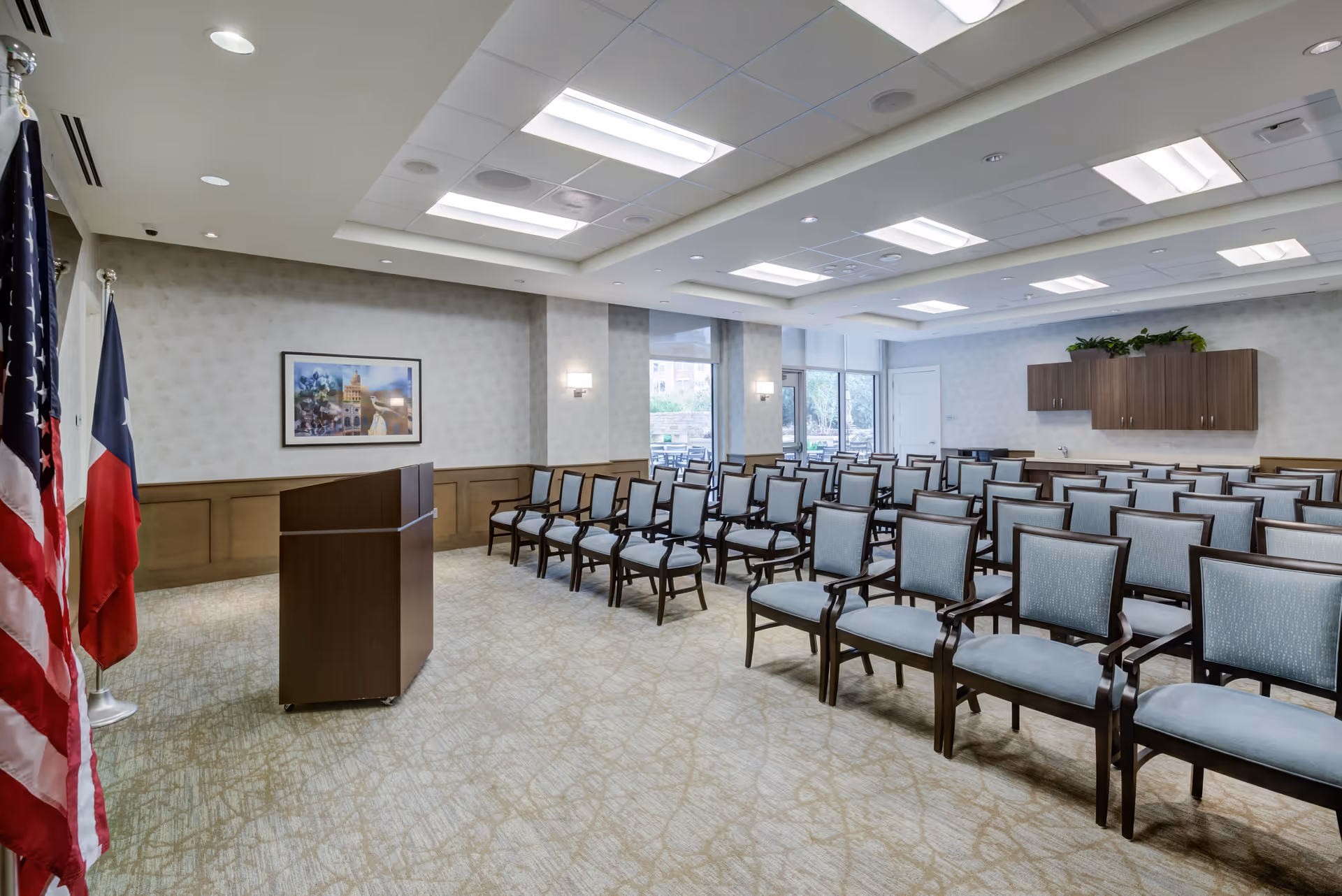 A conference or meeting room with rows of blue upholstered chairs arranged facing a wooden podium. Two flags, one American and one Texas state flag, stand to the left of the podium. The room has beige patterned carpet, light-colored walls with wood paneling, ceiling lights, and large windows letting in natural light. There are wooden cabinets mounted on the far wall with plants on top.