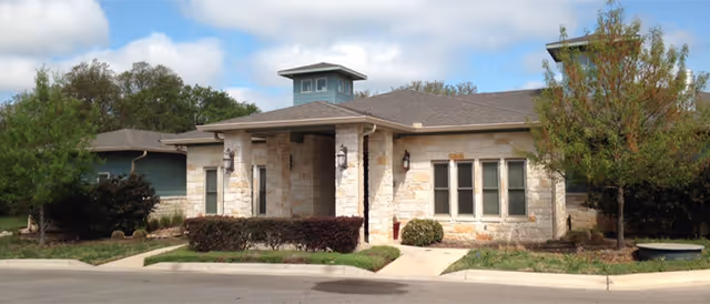 Exterior view of a single-story stone building with a covered entrance, multiple windows, and a small tower-like structure on the roof. The building is surrounded by landscaped bushes, trees, and a paved driveway.