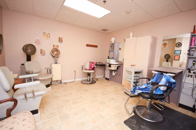 Interior view of a hair salon area with multiple salon chairs, hair dryers, mirrors, and cabinets against light pink walls and a tiled floor.