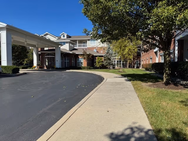 Exterior view of a senior living facility with a covered entrance, paved driveway, sidewalk, green lawn, and trees under a clear blue sky.
