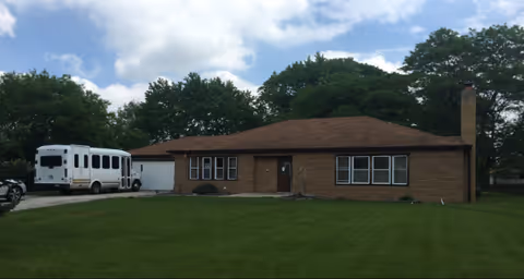 Single-story brick building with a brown roof and multiple windows, surrounded by green grass and trees under a partly cloudy sky. A white shuttle bus is parked on the driveway to the left of the building.