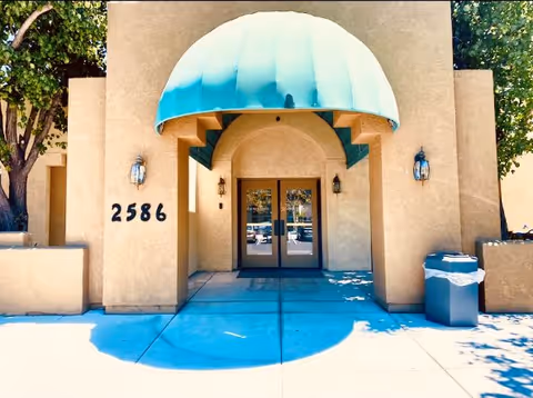 Entrance of a building with the number 2586 displayed on the left wall. The entrance has double glass doors under a large green awning. There are two wall-mounted lantern-style lights on either side of the doors and a trash can on the right side. Trees and shadows are visible around the entrance.
