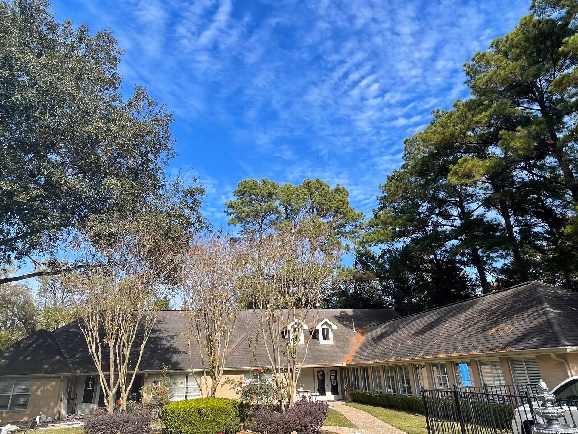 Exterior view of a single-story residential building with a dark shingled roof, surrounded by trees and shrubs under a blue sky with scattered clouds. There is a pathway leading to the entrance, and a parking area with a car visible on the right side.