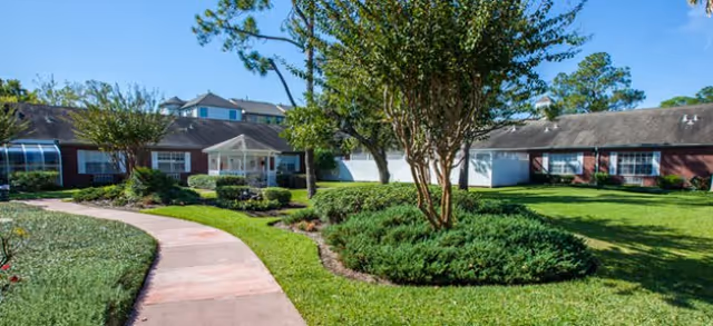 A well-maintained outdoor garden area at Brookdale Memorial Oaks featuring a curved pinkish walkway, green grass, neatly trimmed bushes, and trees. The background shows a single-story building with red brick and white walls under a clear blue sky.