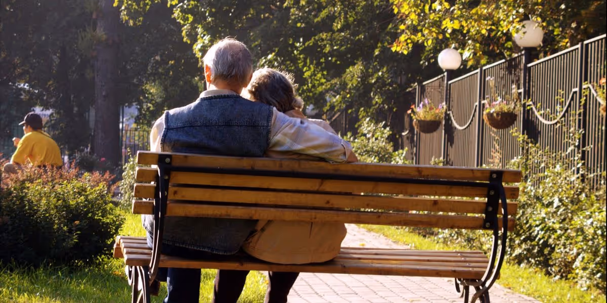 An elderly couple sitting closely together on a wooden bench in a sunlit garden or park area, with greenery and hanging flower pots along a fence in the background.