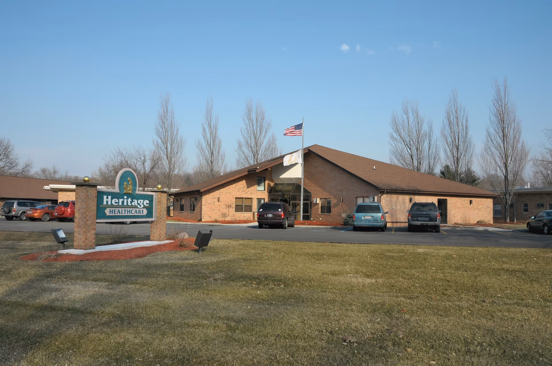 Exterior view of Heritage Healthcare facility showing a single-story brick building with a brown roof, an American flag on a flagpole in front, several parked cars, and a green sign with the facility name on a lawn under a clear blue sky.