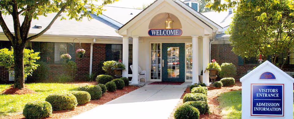 Front entrance of a brick building with a covered porch and a 'WELCOME' sign above the door. The walkway is lined with neatly trimmed bushes and trees. A sign near the entrance reads 'VISITORS ENTRANCE ADMISSION INFORMATION'.