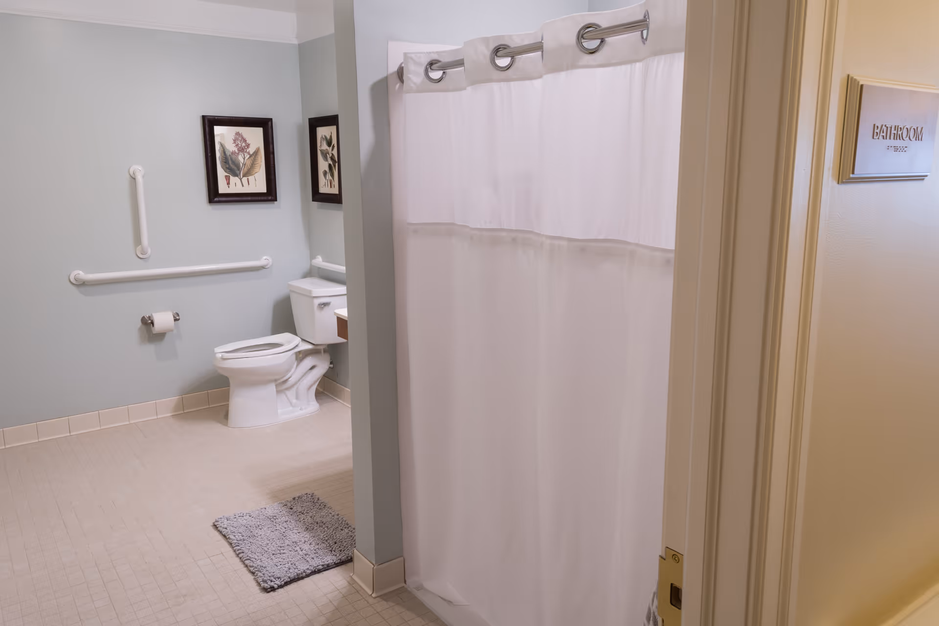 A clean and accessible bathroom with a white toilet, grab bars on the wall, a toilet paper holder, a small gray rug on the tiled floor, two framed botanical pictures on the wall, and a white shower curtain partially visible. The door has a sign that reads 'BATHROOM'.