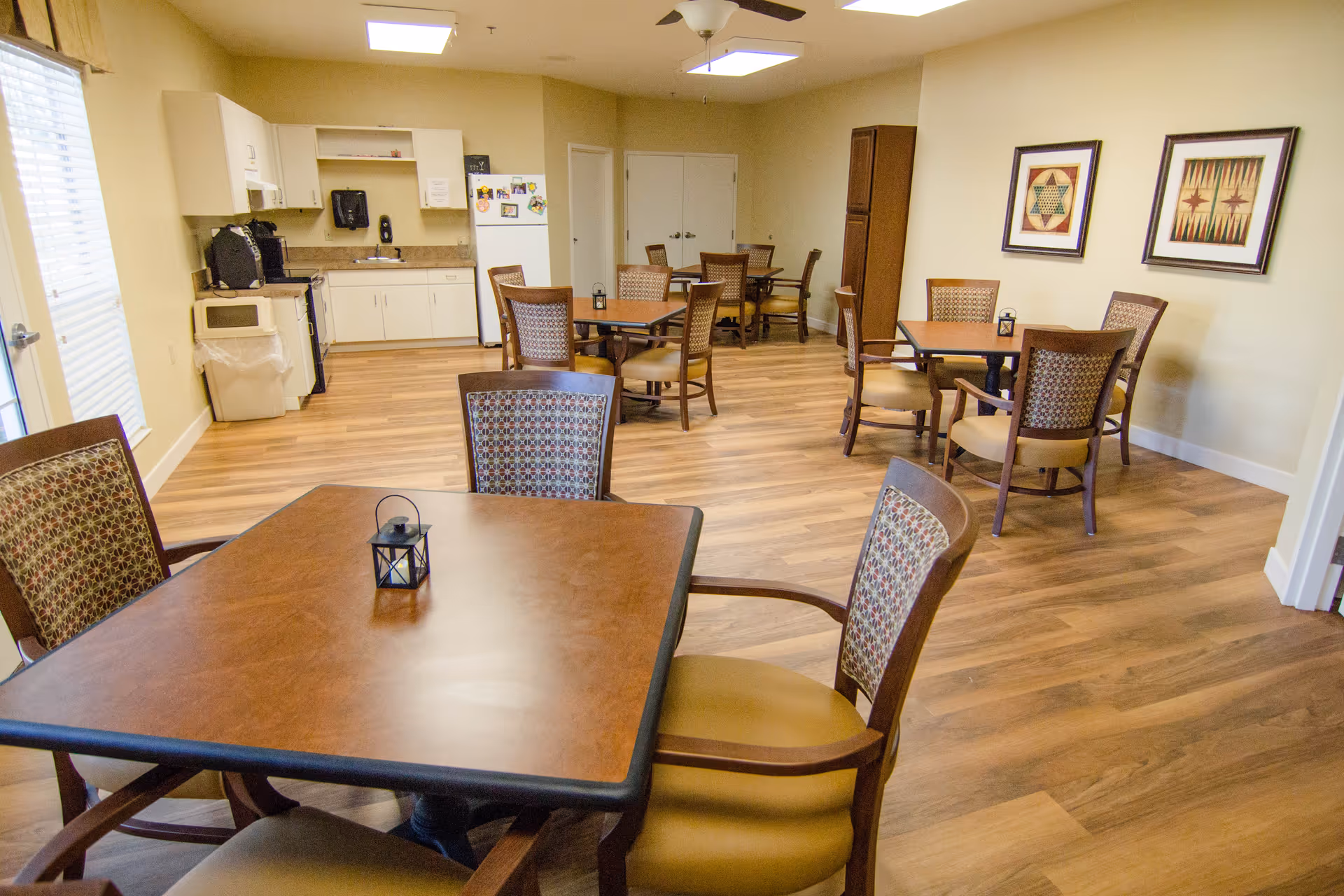 A communal dining area with several square wooden tables and cushioned chairs arranged around the room. The room has light-colored walls, wood flooring, and a ceiling fan with lights. In the background, there is a small kitchen area with white cabinets, a refrigerator, a microwave, and a coffee maker. Two framed artworks hang on the wall to the right.