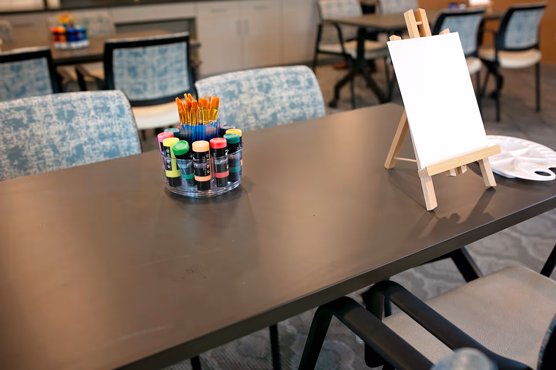 A table in a communal room with a small easel holding a blank canvas, a paint palette, and a container filled with paintbrushes and colorful paint bottles. Chairs with patterned upholstery surround the table, and more tables and chairs are visible in the background.