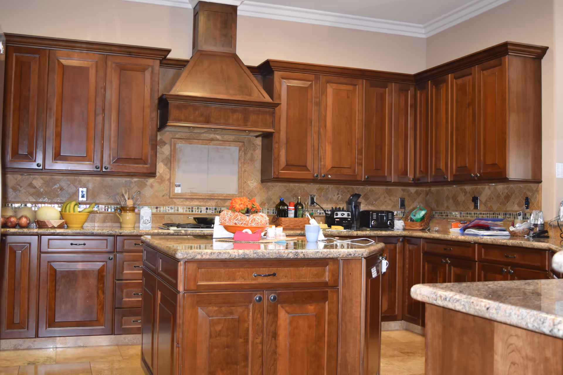 A kitchen with wooden cabinets and drawers, a central island with a granite countertop, and various kitchen items including a bowl of bananas, a toaster, a coffee maker, and some decorative items on the counter.