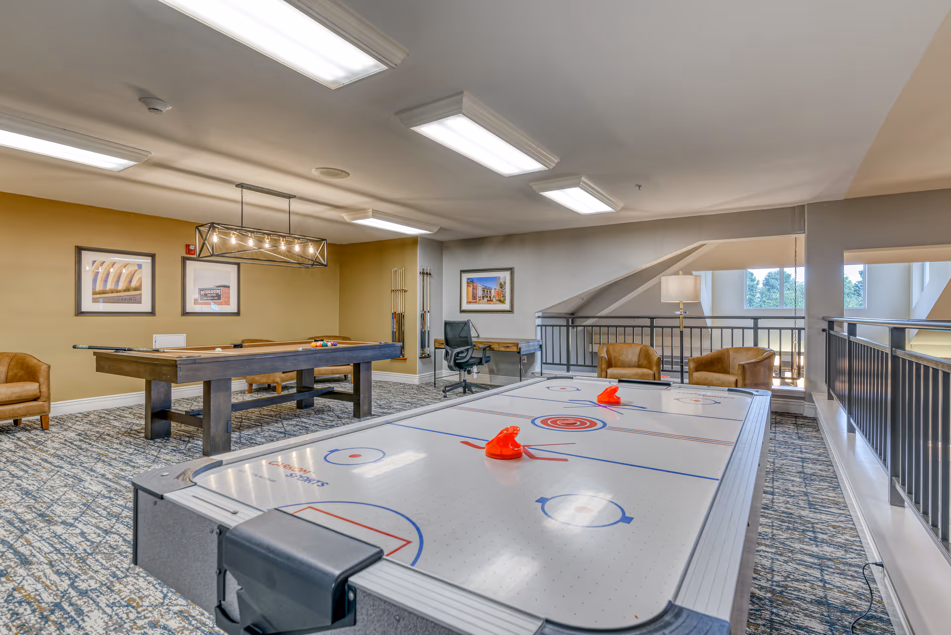 A recreational room featuring an air hockey table in the foreground and a pool table in the background. The room has carpeted flooring, beige walls, and several framed pictures on the walls. There are two brown armchairs near a railing overlooking a lower level, and a desk with an office chair is positioned against the far wall. The room is well-lit with ceiling lights and a hanging light fixture above the pool table.