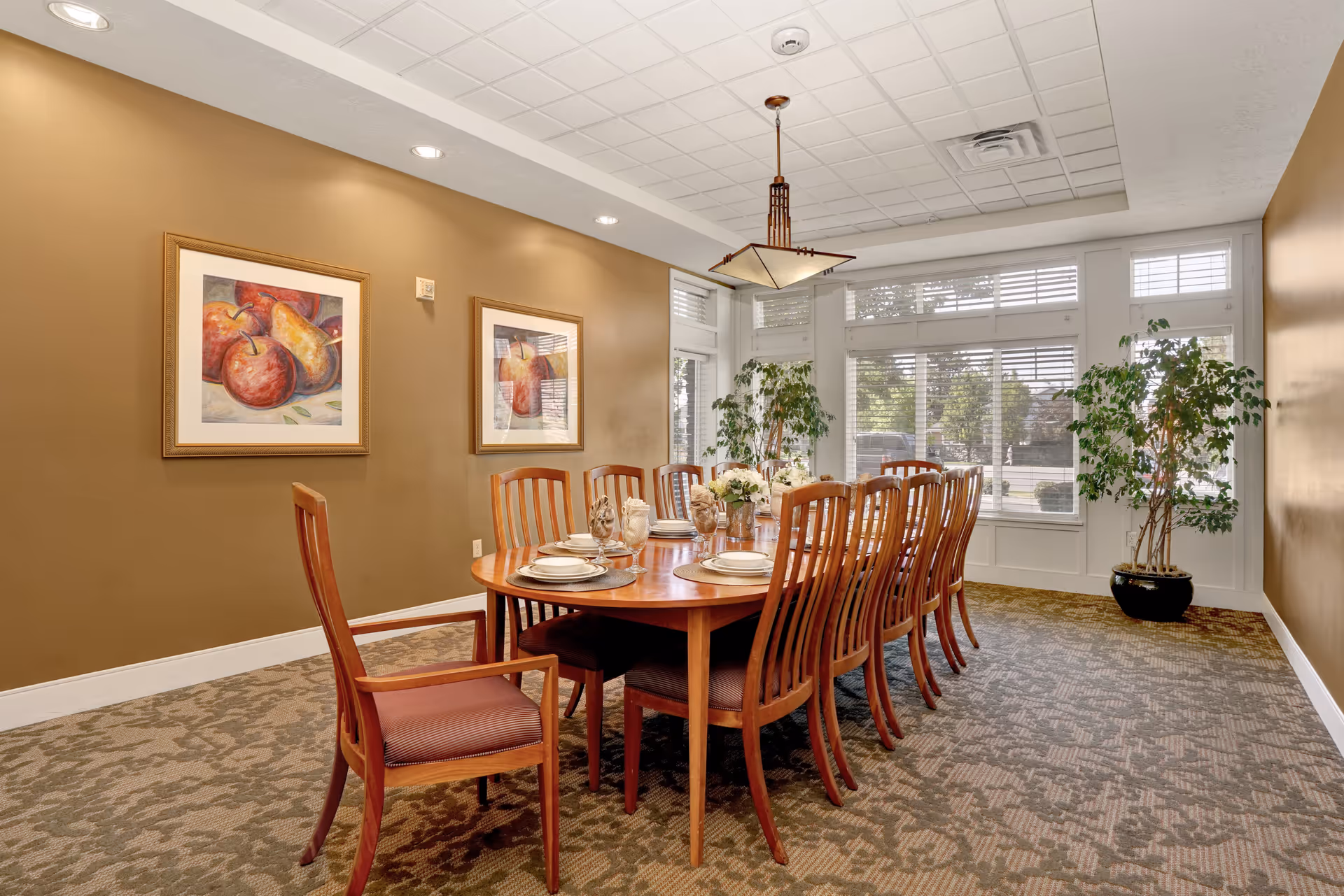 A well-lit dining room with a long wooden table set for ten people. The table is adorned with plates, glasses, and floral centerpieces. The room has beige walls with two framed paintings of fruit, large windows with white blinds, and two potted plants near the windows. A decorative ceiling light fixture hangs above the table.