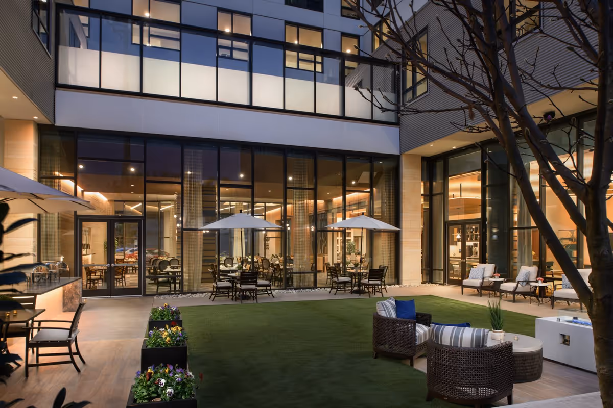 Outdoor courtyard area at dusk with seating arrangements including wicker chairs with cushions, tables, umbrellas, and planters with flowers. Large glass windows and doors reveal an indoor dining area with tables and chairs inside a modern building.