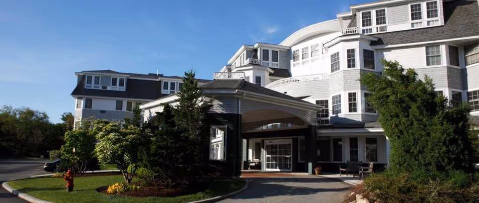 Exterior view of a large, multi-story senior living facility building with white siding and numerous windows under a clear blue sky. The entrance features a covered driveway with greenery and trees surrounding the area.
