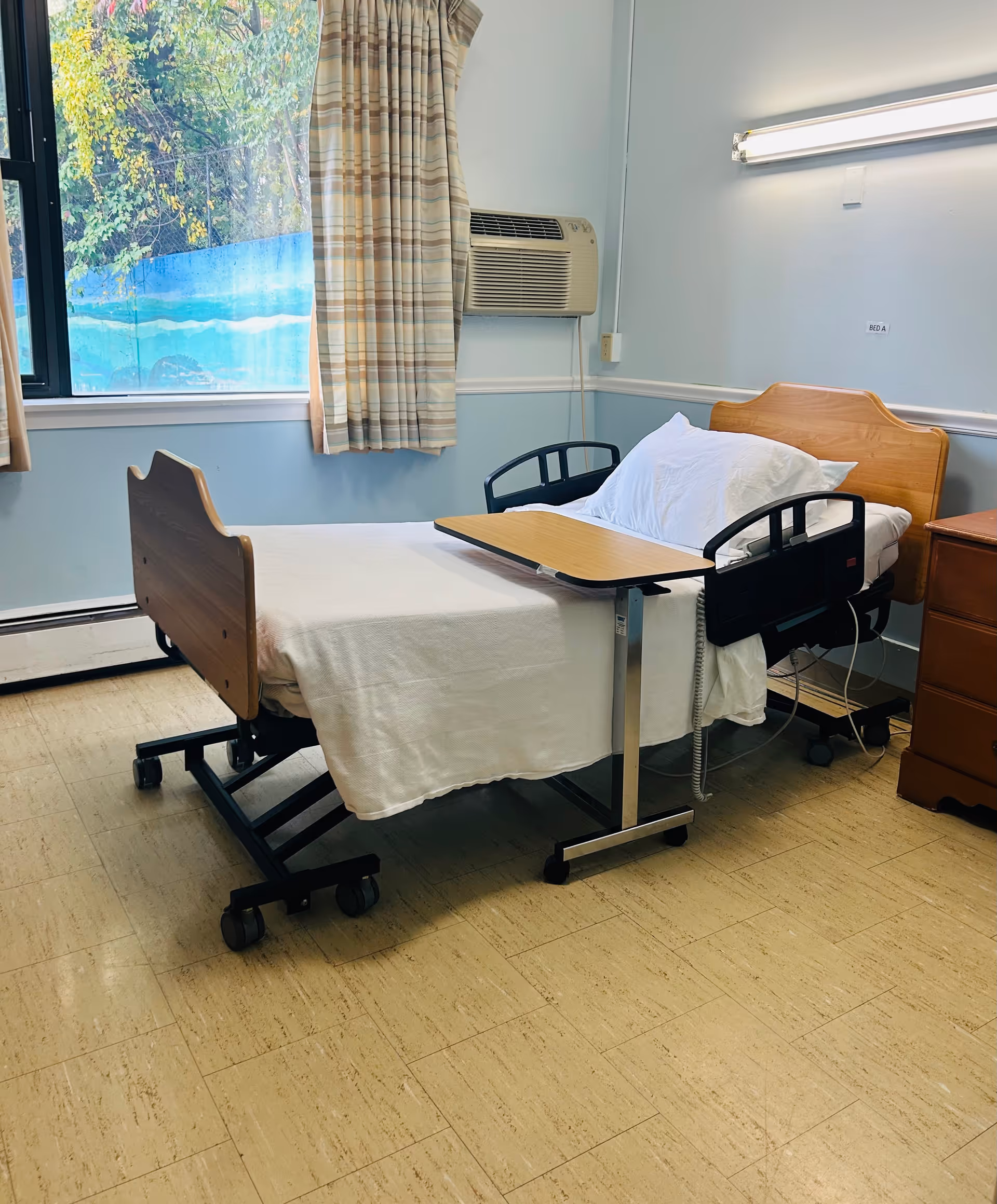 A hospital-style bed with white sheets and pillows in a room with light blue walls. The bed has black side rails and an overbed table. There is a window with beige and brown striped curtains, an air conditioning unit mounted on the wall, and a wooden bedside cabinet. The floor is covered with beige tiles.