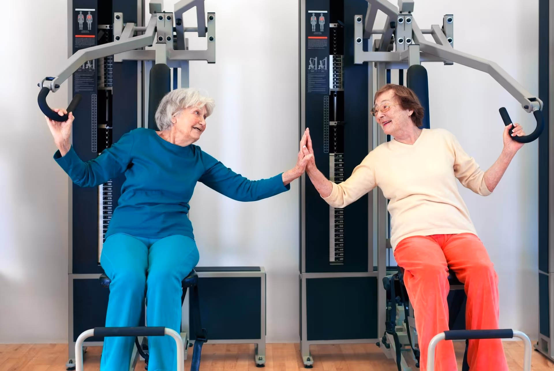 Two elderly women sitting on exercise machines in a fitness room, smiling and giving each other a high-five while holding the machine handles.