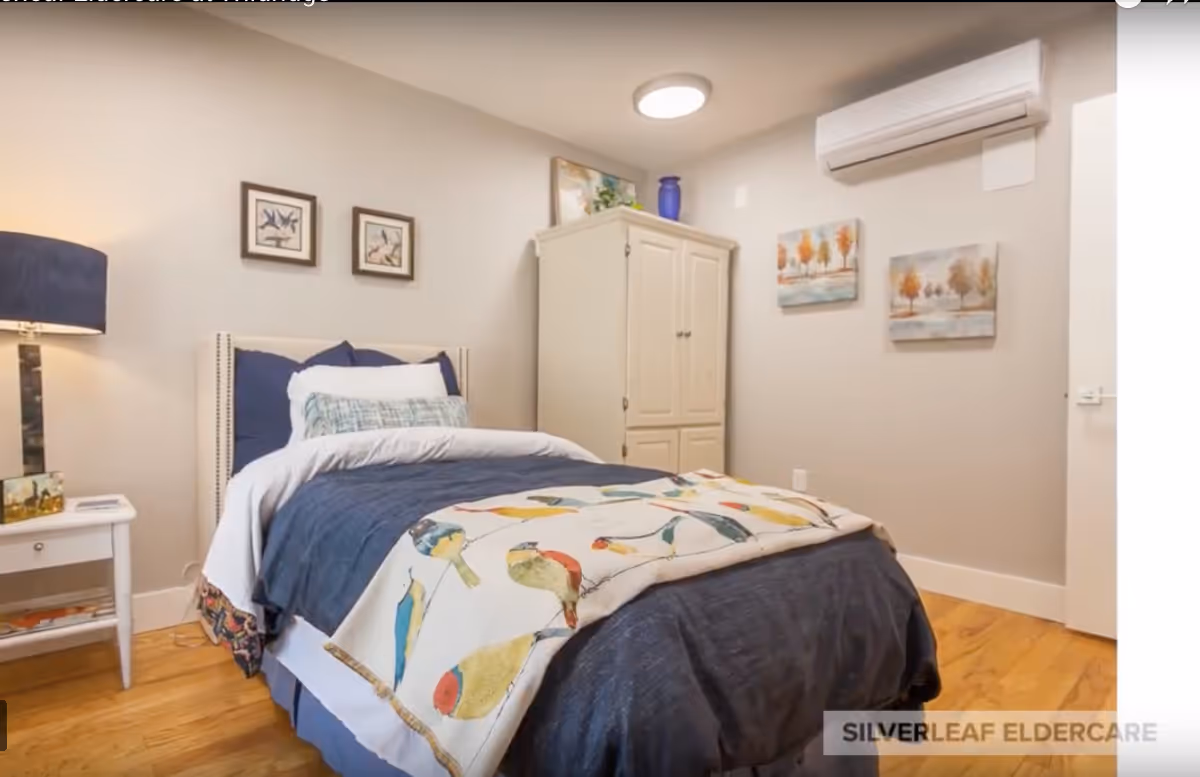 A cozy bedroom with a single bed dressed in blue bedding and a colorful bird-patterned blanket. There is a white nightstand with a lamp and some items on it to the left of the bed. On the wall above the bed are two framed bird illustrations. A white wardrobe stands against the far wall, with two paintings of autumn trees hanging nearby. The room has wooden flooring and a wall-mounted air conditioning unit.