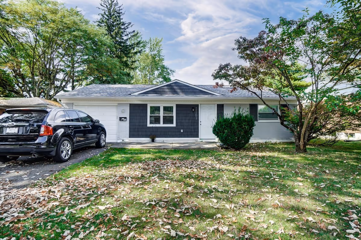 Single-story house with a dark gray and white exterior, a white front door, and a garage. A black SUV is parked in the driveway. The front yard has green grass with scattered fallen leaves and several trees around the property under a partly cloudy sky.