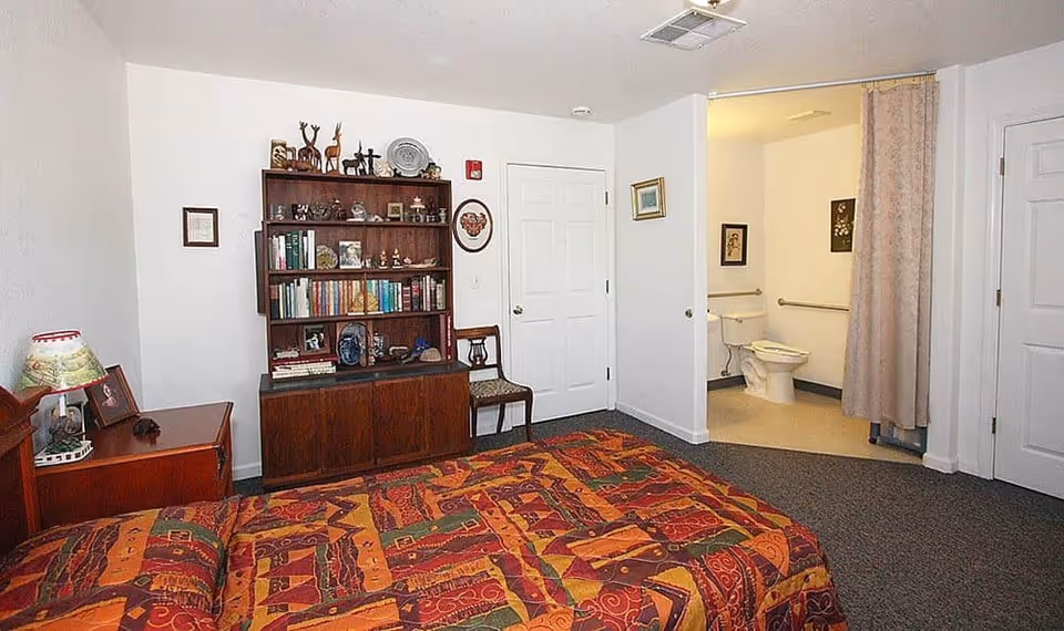 A bedroom in Oak Terrace Memory Care featuring a bed with a colorful patterned bedspread, a wooden nightstand with a lamp and framed photo, a wooden bookshelf filled with books and decorative items, a chair, and an open doorway leading to a bathroom with a toilet and grab bars. The walls are white and the floor is carpeted.