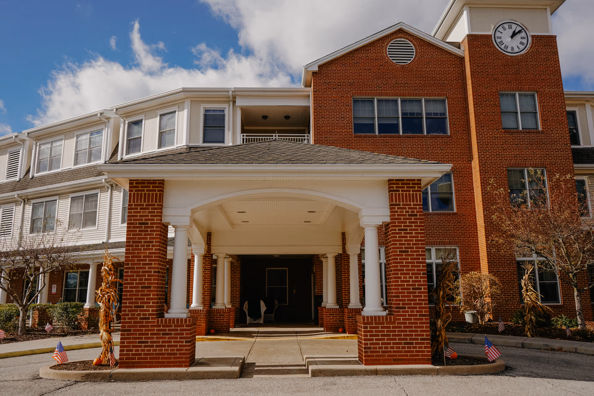 Front exterior view of a senior living facility building with a covered entrance supported by brick columns and white pillars. The building has a clock tower on the right side and multiple windows. There are small American flags and autumn decorations near the entrance.