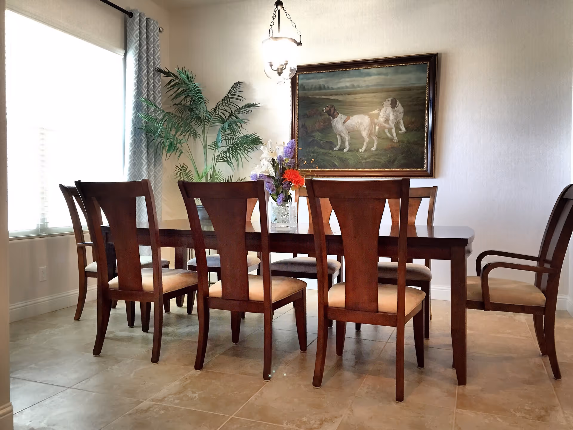 A dining room with a wooden dining table and eight matching wooden chairs with beige cushions. A vase with colorful flowers is placed on the table. There is a large painting of two dogs on the wall and a tall green plant near a window with patterned curtains. The floor is tiled and a hanging light fixture is above the table.