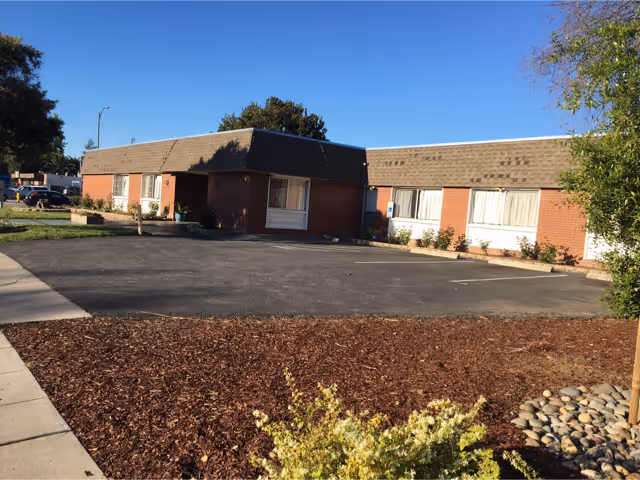 Exterior view of a single-story brick building with multiple windows, a shingled roof, and a parking area in front. There are some small bushes and landscaped areas with mulch and rocks near the building. The sky is clear and blue.