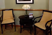 Two beige upholstered chairs with dark wooden frames are positioned on either side of a small round dark wooden table. On the table, there is a vase with white flowers and a small brochure or card. The background wall is light green with a wooden chair rail and a framed picture hanging above.