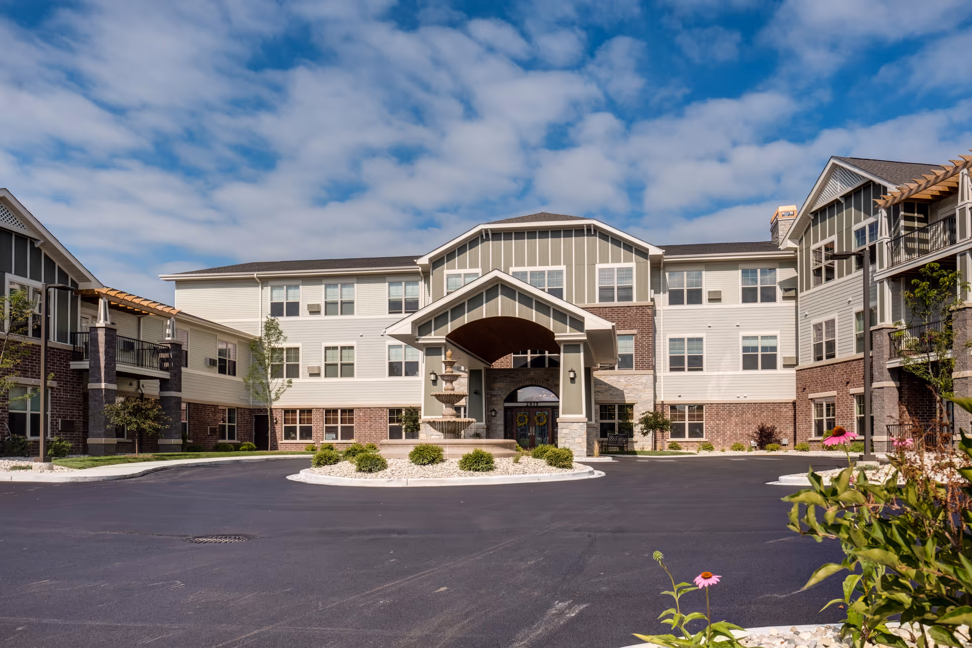 Front exterior view of a large senior living facility building with a covered entrance, a three-tiered fountain in the circular driveway, landscaped bushes, and flowers under a partly cloudy blue sky.