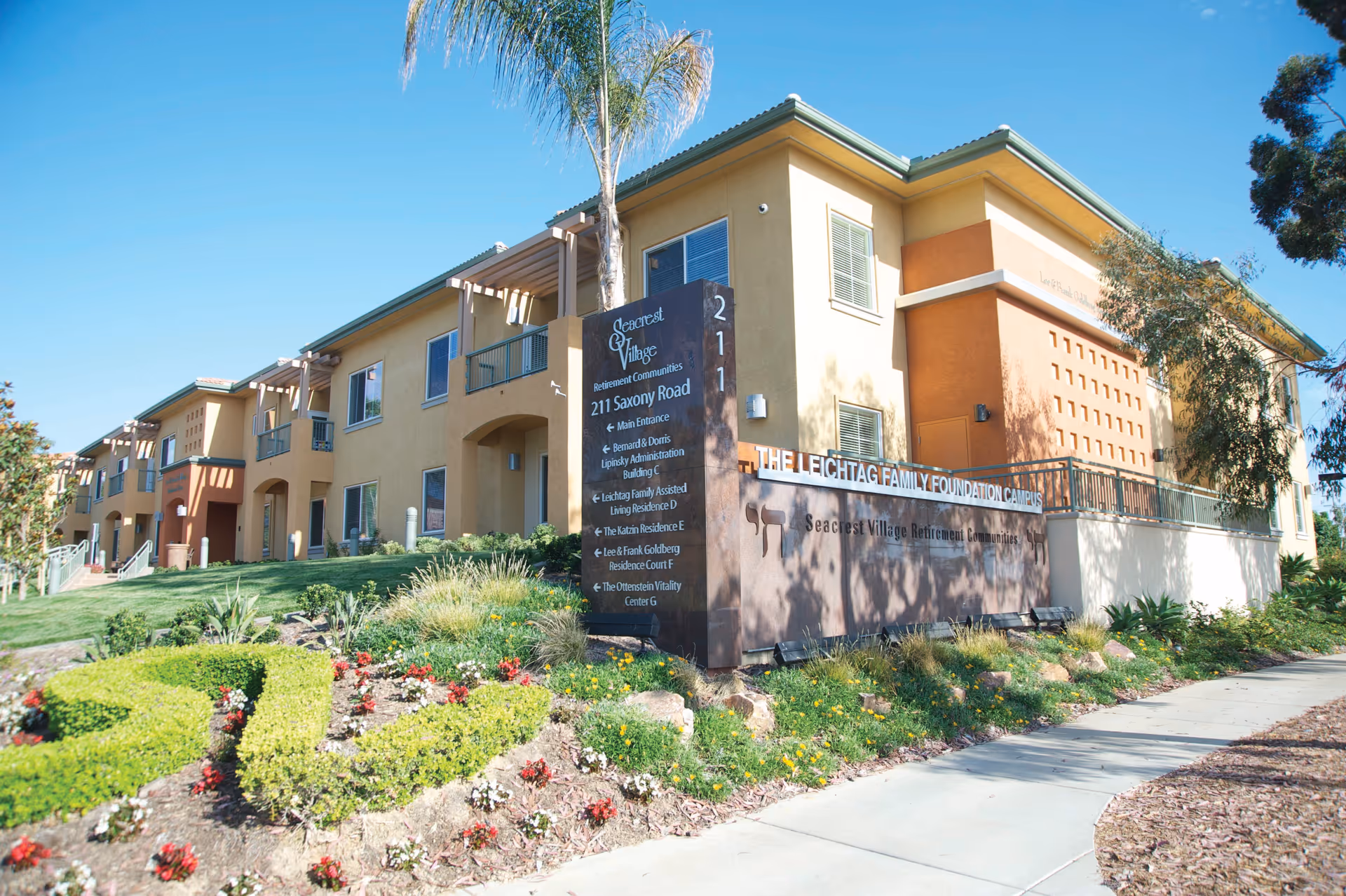 Exterior view of Seacrest Village Retirement Communities building with a landscaped garden and a large sign displaying the facility's name and address at 211 Saxony Road under a clear blue sky.