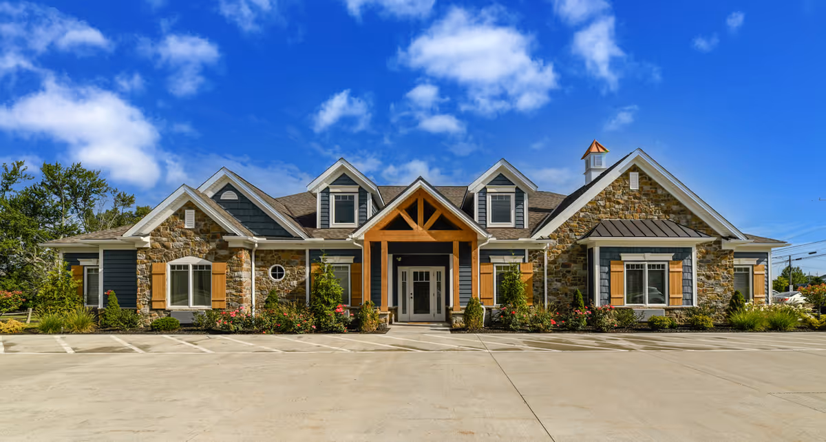 Front exterior view of a single-story building with stone and wood siding, multiple gabled roofs, and a covered entrance supported by wooden beams. The building is surrounded by landscaped bushes and flowers, with a clear blue sky above and a concrete parking area in front.