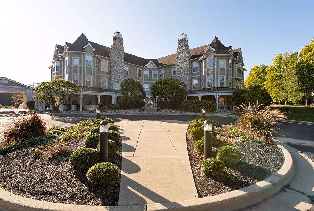 Front exterior view of a large, multi-story senior living facility named Bloom at Kessler, featuring stone chimneys, multiple windows, a covered porch, and landscaped garden beds with shrubs and ornamental grasses along a paved driveway.