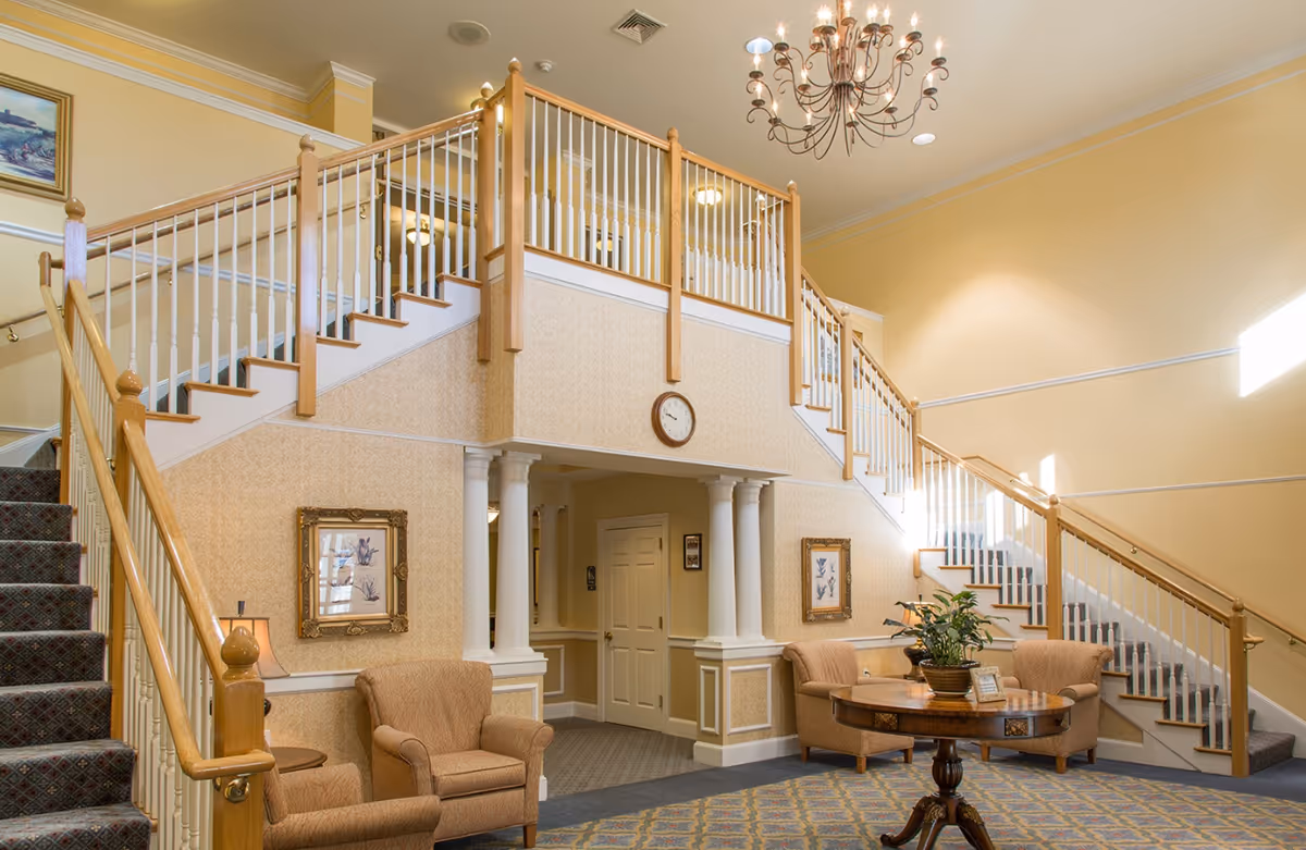 Interior view of a senior living facility lobby area with a staircase leading to an upper floor. The space features beige walls, a chandelier, two armchairs, framed artwork, a round wooden table with a plant, and a clock mounted on the wall above a doorway.