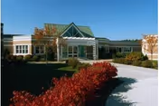 Exterior view of Schuyler Ridge Residential Healthcare building with a clear blue sky, landscaped bushes with red leaves in the foreground, and a paved walkway leading to the entrance.