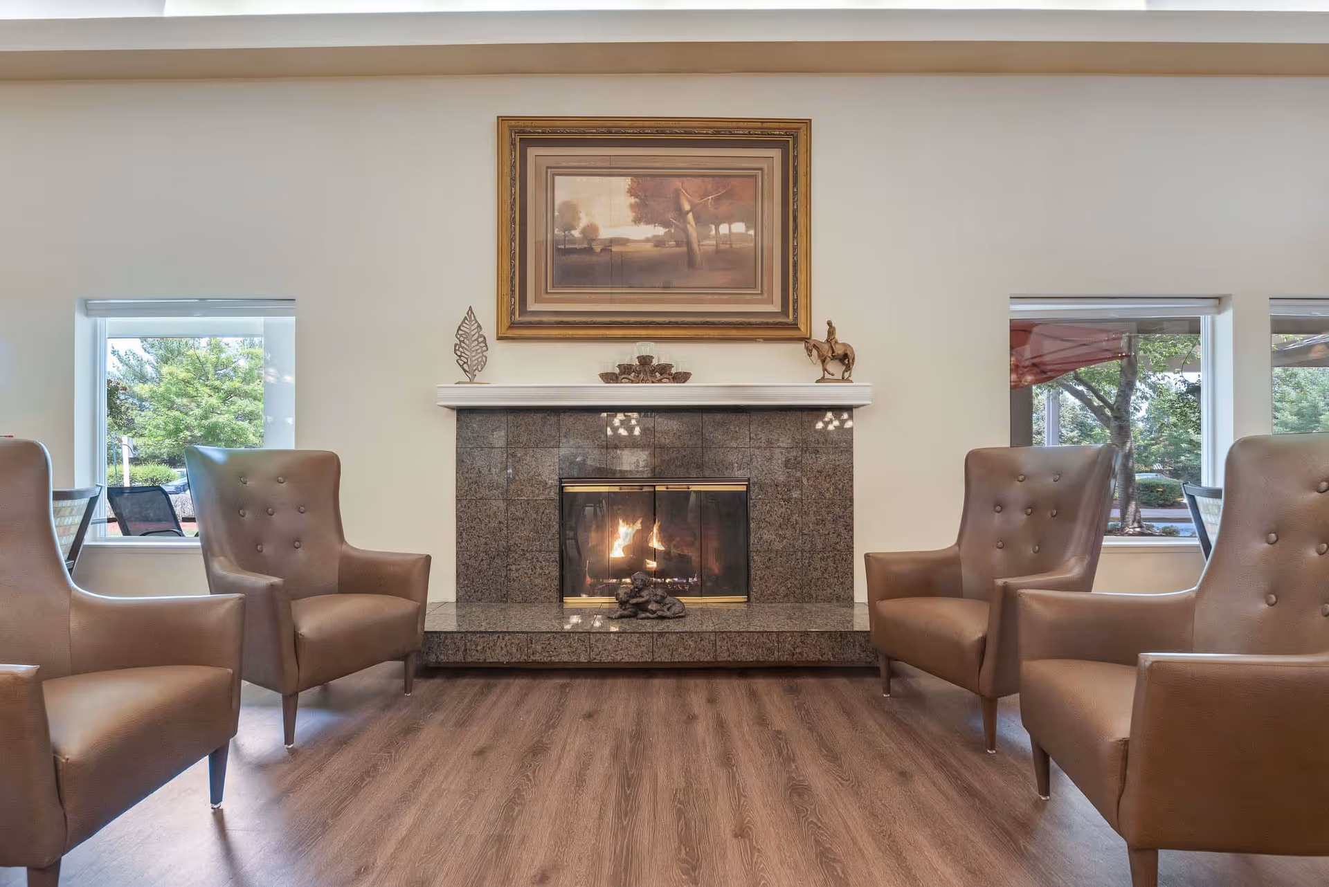 A cozy living room area with a lit fireplace surrounded by dark tiles. Four brown leather armchairs are arranged facing the fireplace. Above the fireplace is a framed painting of a tree landscape, and decorative items are placed on the mantel. Two windows on either side of the fireplace show greenery and outdoor seating.