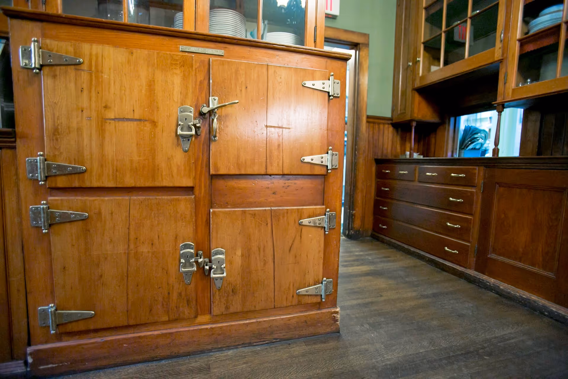 A close-up view of a vintage wooden icebox with metal hinges and latches in a room with wooden cabinetry and drawers, featuring plates and bowls stored in glass-front cabinets above.