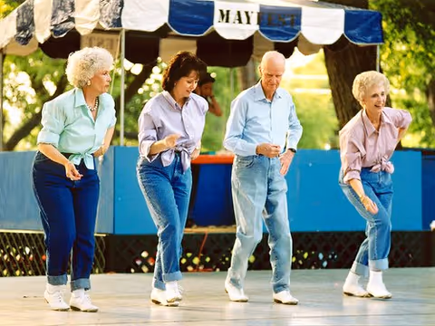 Four elderly people line dancing outdoors on a wooden stage under a blue and white striped canopy with the word 'MAYFEST' on it. They are dressed casually in jeans and light-colored shirts, enjoying the activity together.